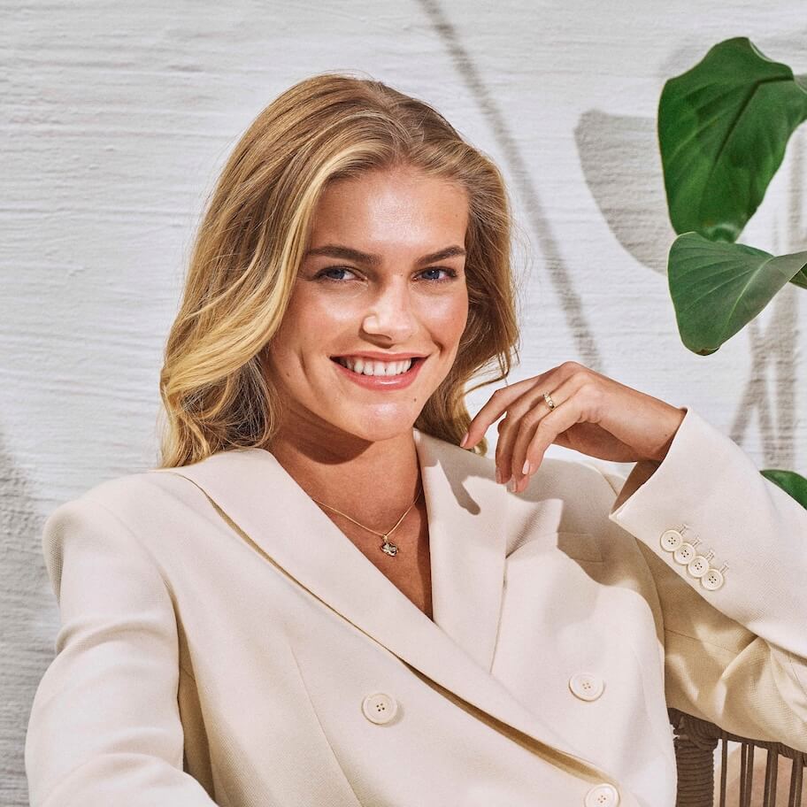 Smilende kvinne med langt blondt hår i en kremfarget blazer, sittende i nærheten av en grønn plante mot en lys teksturert bakgrunn.